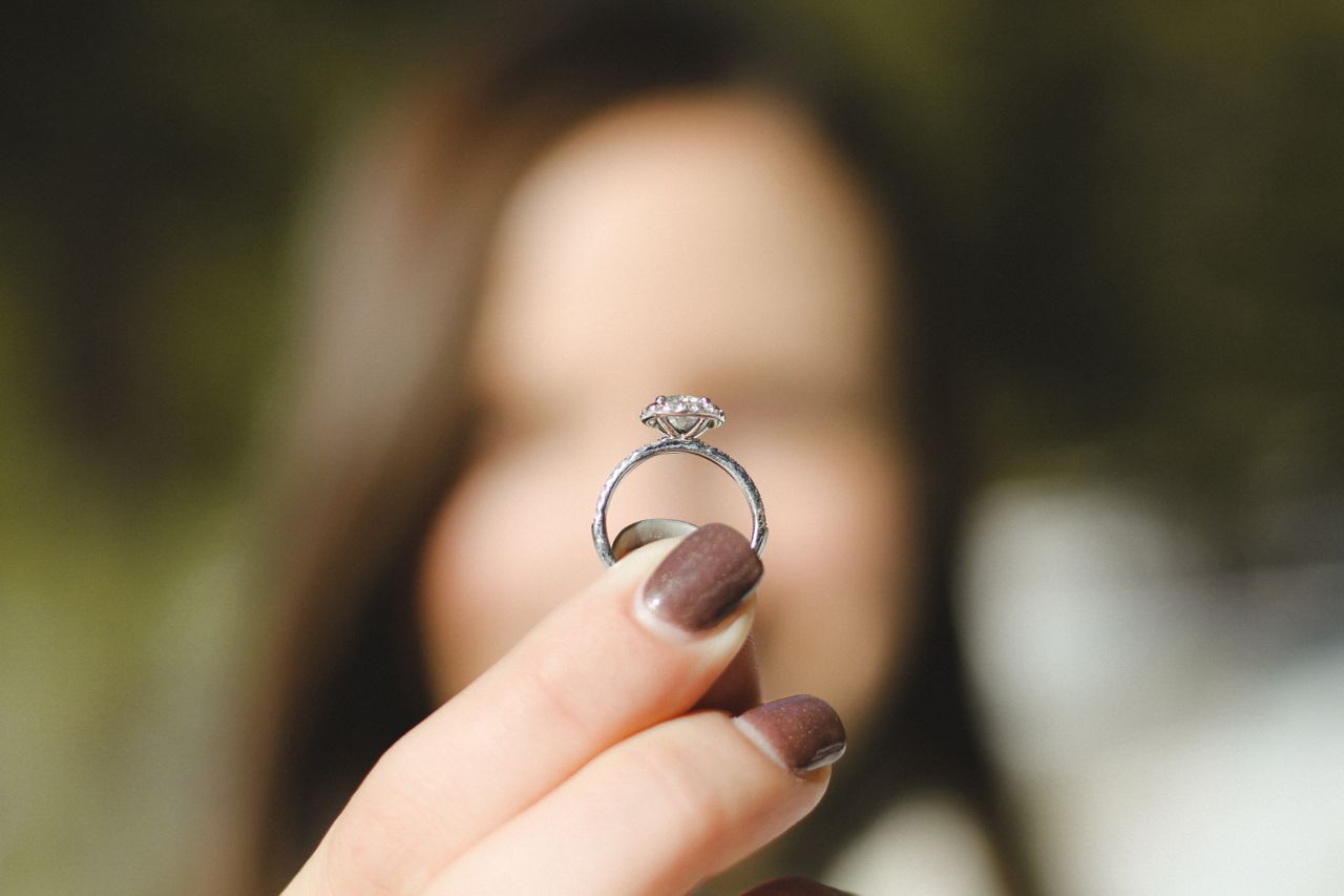 Solitaire engagement ring held up to the camera by a fuzzy individual.