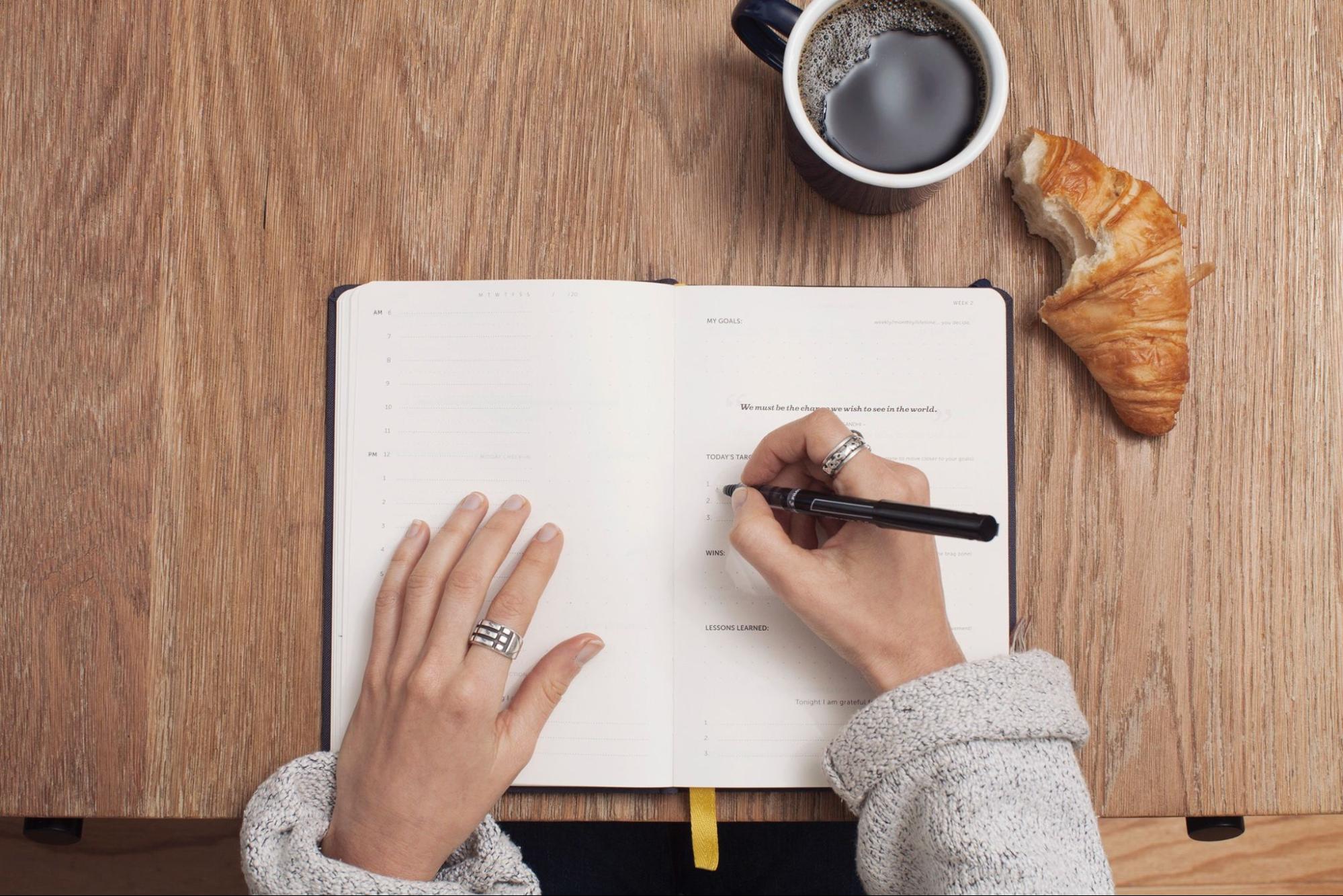 An overhead view of a woman writing in a planner, her hands adorned with two rings.