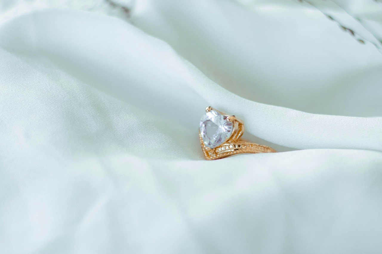 A delicate gold engagement ring with a heart shaped center stone, carefully displayed within the folds of sheer white fabric.