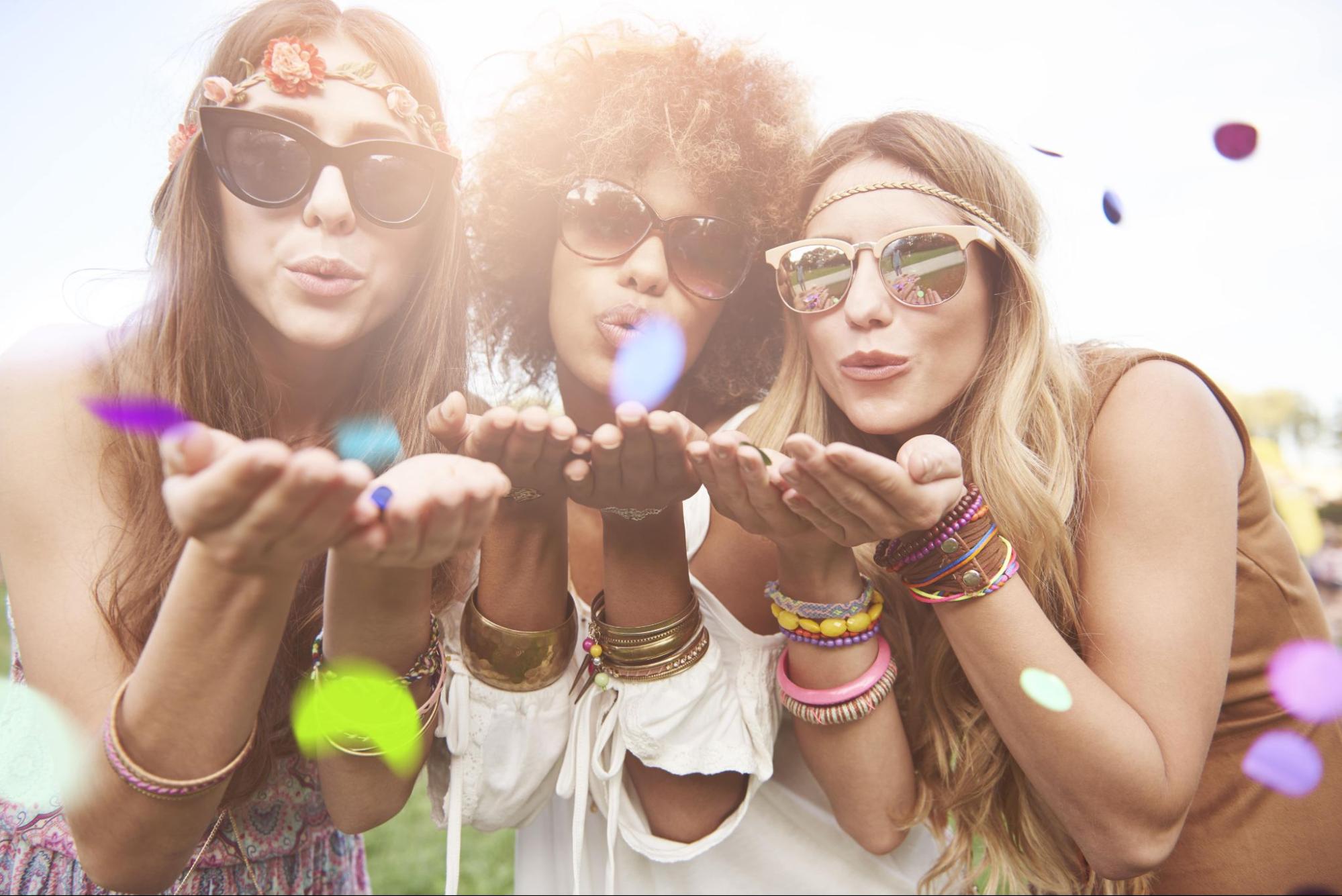 Three bridesmaids adorned in sunglasses and many colorful bracelets blowing confetti toward the camera.