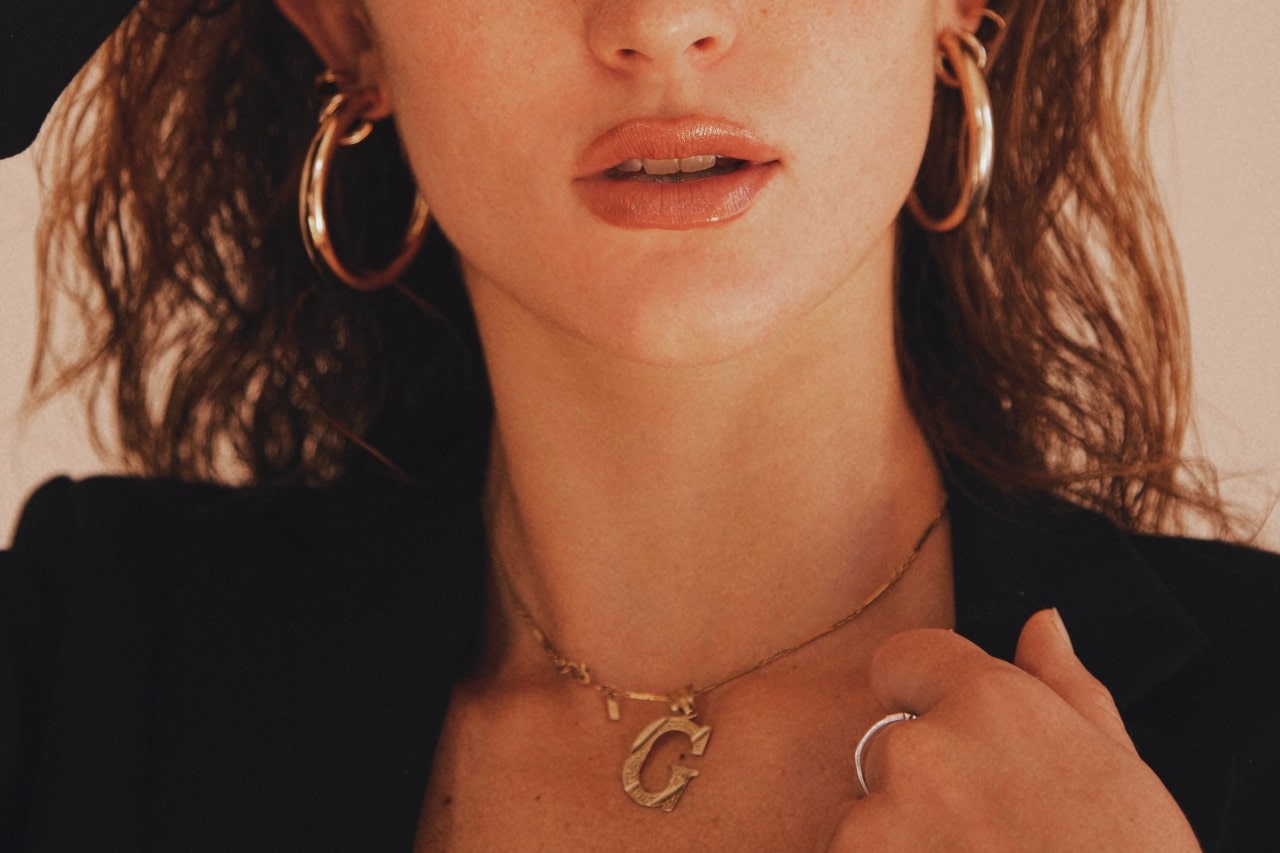 A woman wearing yellow gold hoop earrings a matching “g” pendant necklace along with a white gold ring