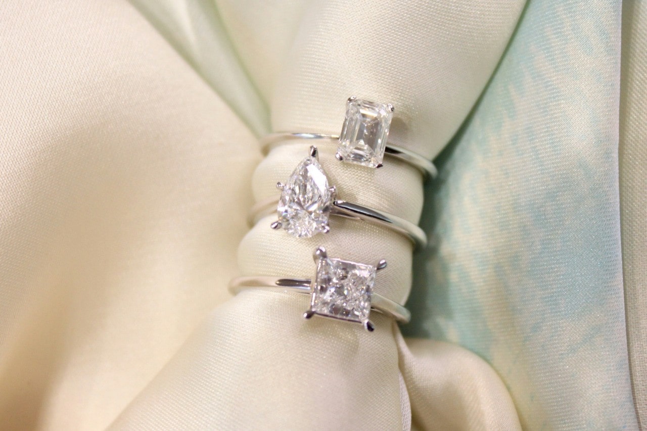 A close-up of three solitaire rings featuring different diamond cuts, displayed on an off-white cloth.