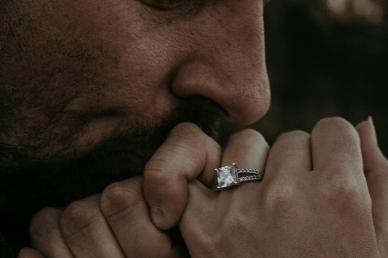 a man kissing a woman’s hand that is wearing a princess cut engagement ring