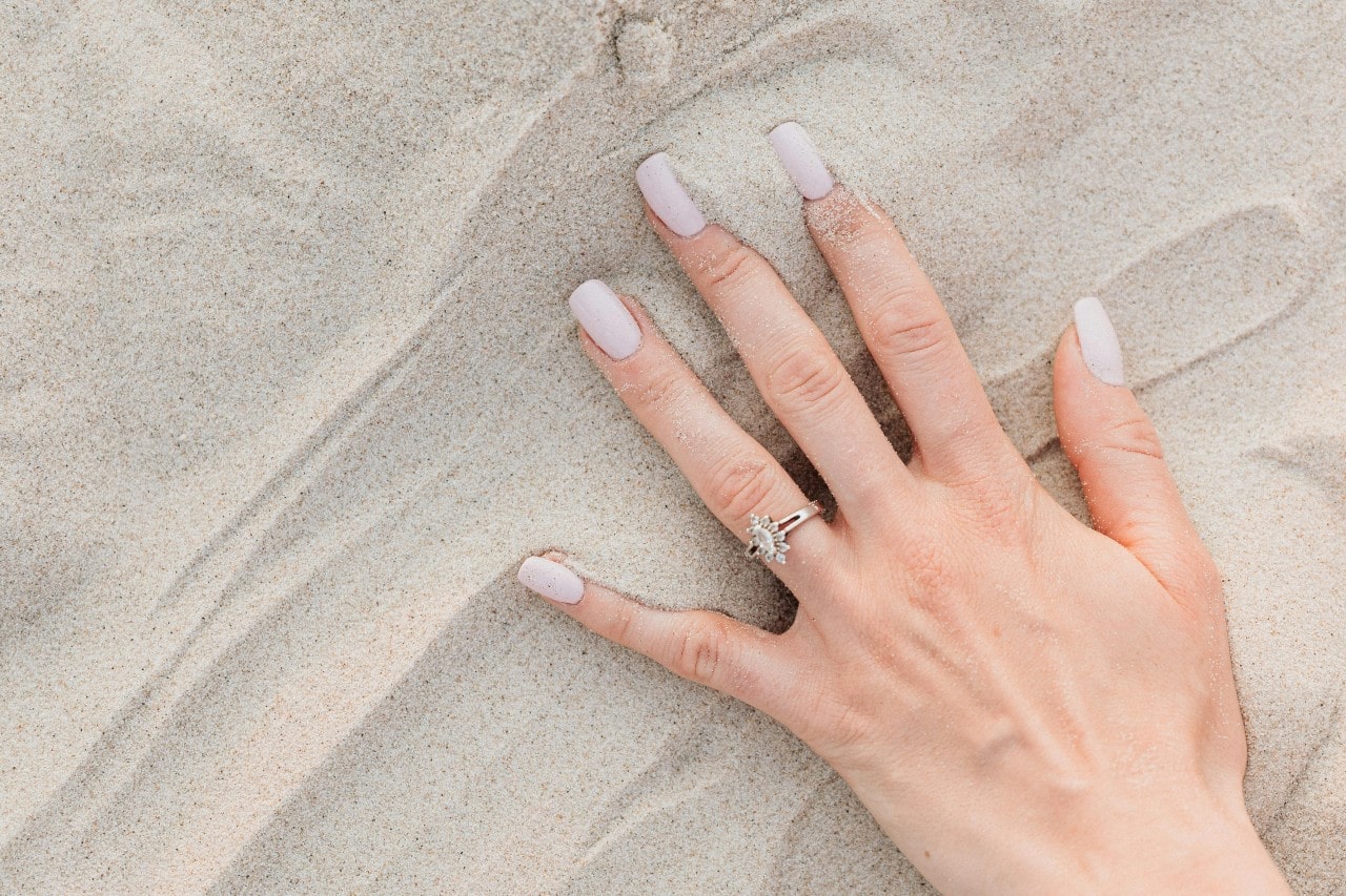 a woman’s hand in the sand, wearing a halo marquise engagement ring