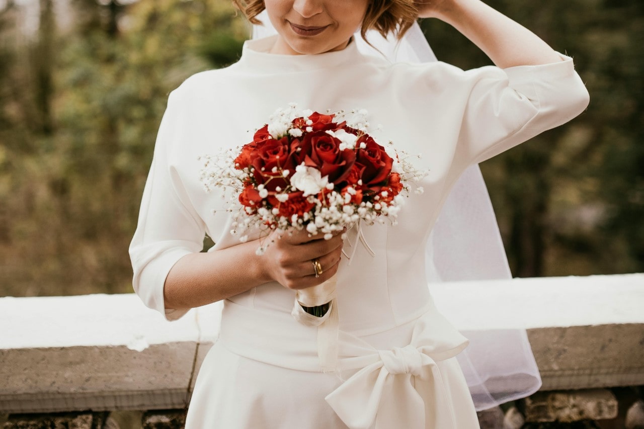 A bride standing outside and see her wedding band and engagement ring holding onto her bouquet of red roses