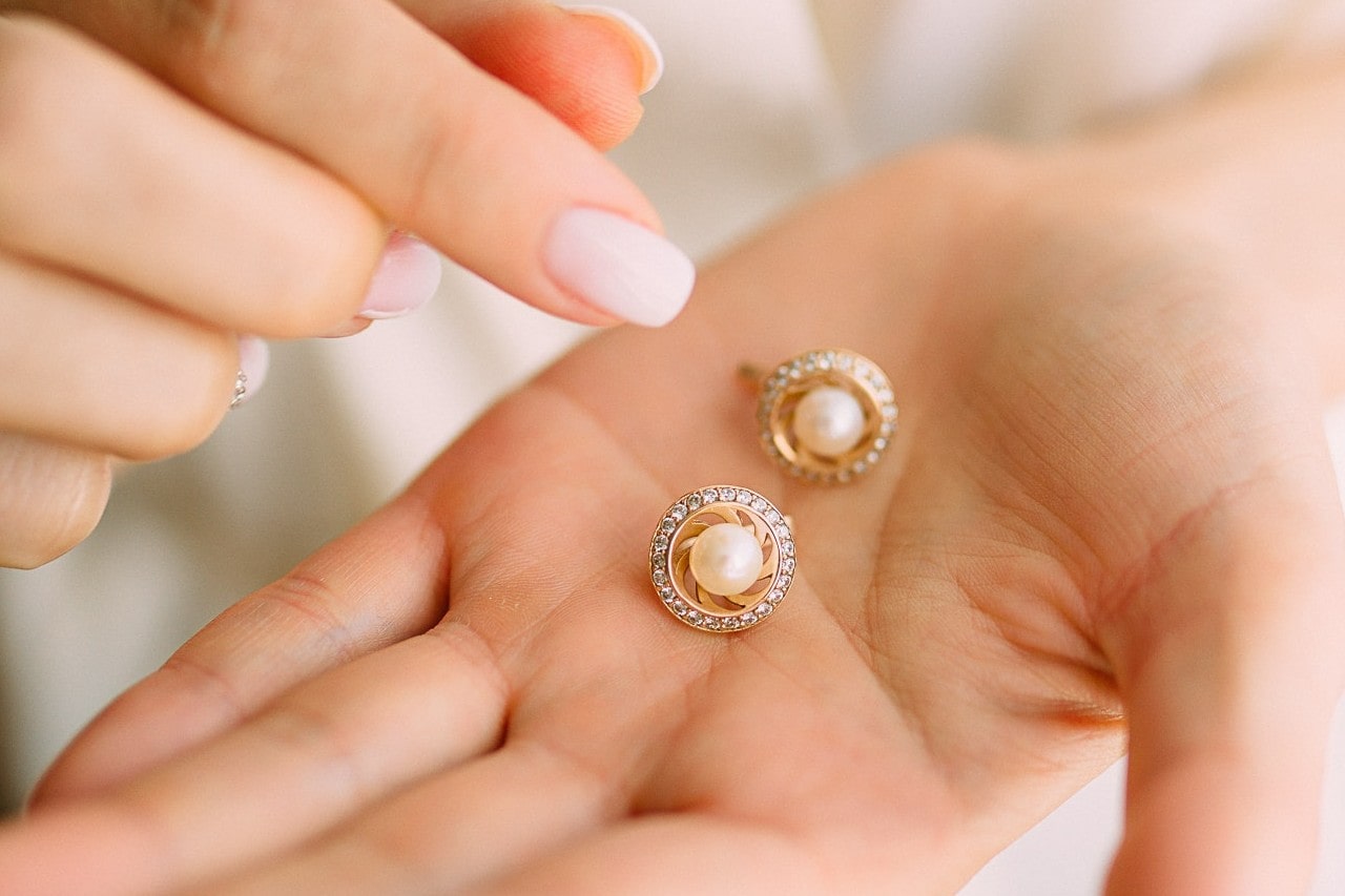 A close-up of a woman's hand holding a pair of round gold earrings, each featuring a central white pearl surrounded by a circle of small sparkling diamonds