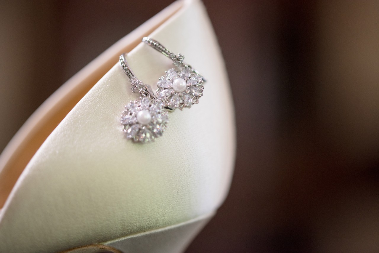 A pair of elegant silver earrings adorned with small, clear gemstones and centered with a white pearl, delicately placed on the pointed toe of an ivory satin high heel shoe.