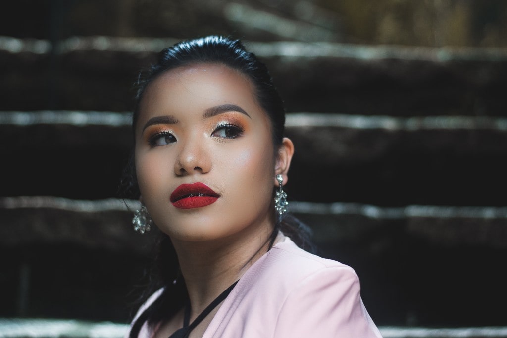 A woman with bold red lipstick and shimmering earrings gazes into the distance, wearing a soft pink blazer against a dark, blurred staircase background.