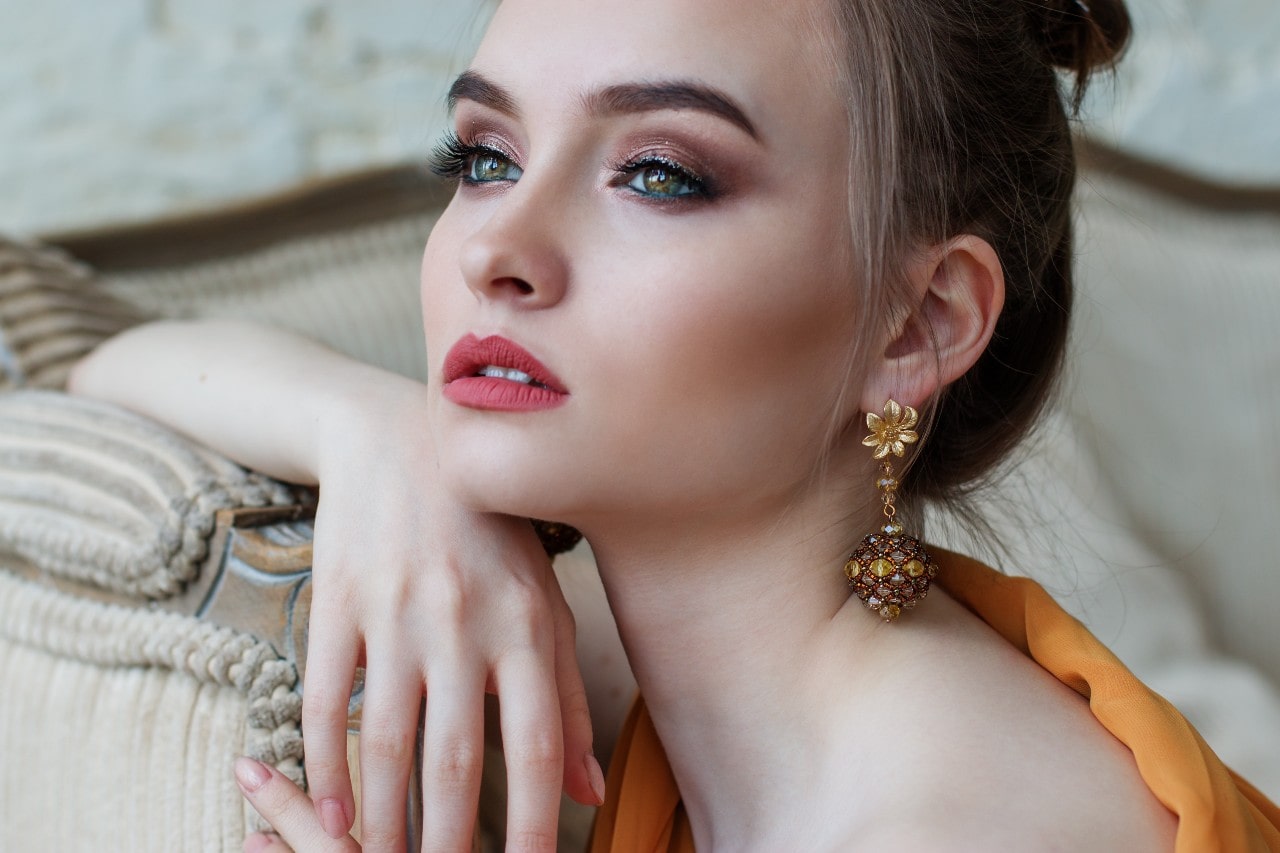 A woman with striking makeup and bold lipstick wearing ornate, dangling gold earrings with a floral design.